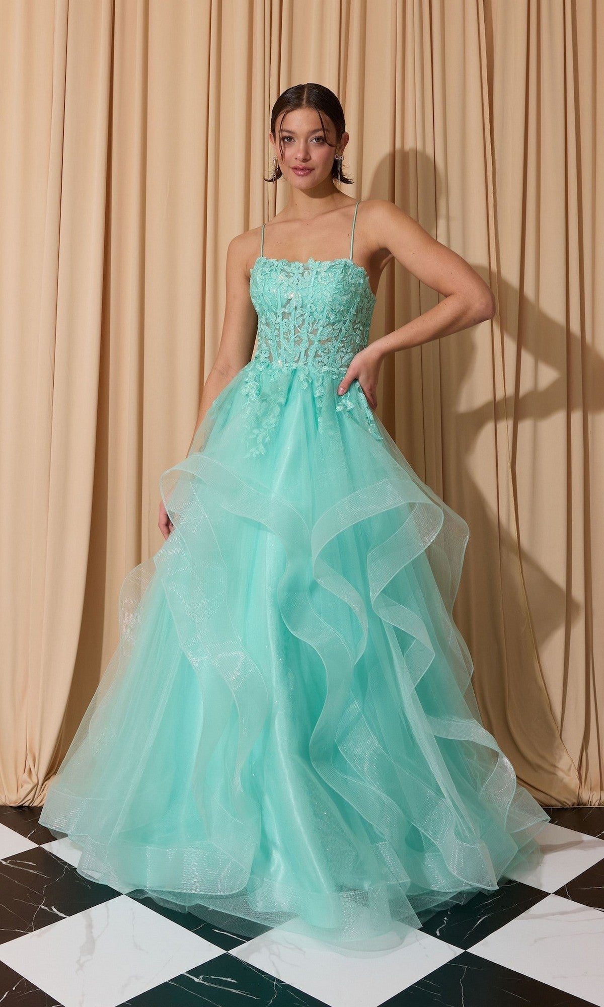 A woman in the Eureka EK167 long prom dress in mint, featuring a floral lace embroidered corset and layered tulle A-line skirt, stands before beige curtains on a black-and-white tiled floor.